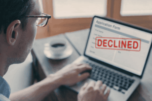 A business owner looking at his laptop screen, which shows an application form with a red 'DECLINED' stamp, illustrating a trademark rejected by the USPTO.